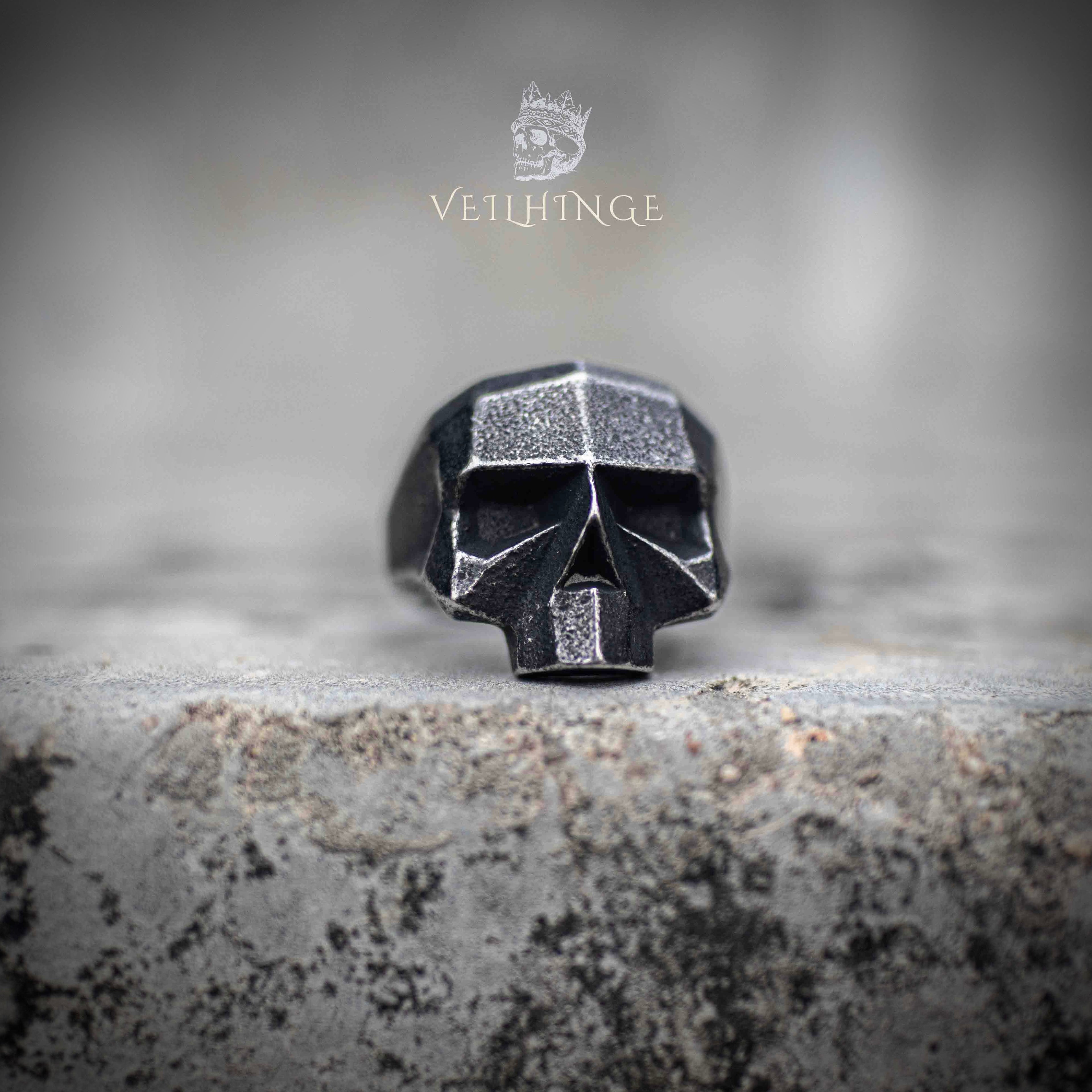 Side profile of geometric skull ring showing oxidized exterior and brushed steel inner band — angular low-poly construction on stone surface