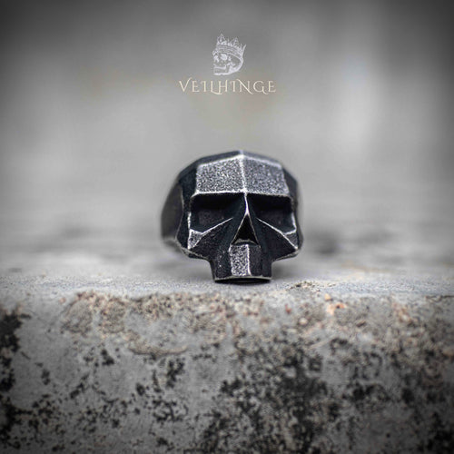 Side profile of geometric skull ring showing oxidized exterior and brushed steel inner band — angular low-poly construction on stone surface