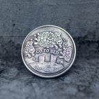 Close-up of the Go To Bed face of a coin engraved with woolly sheep jumping over a moonlit hill, stars and ZZZ symbols, on dark stone surface