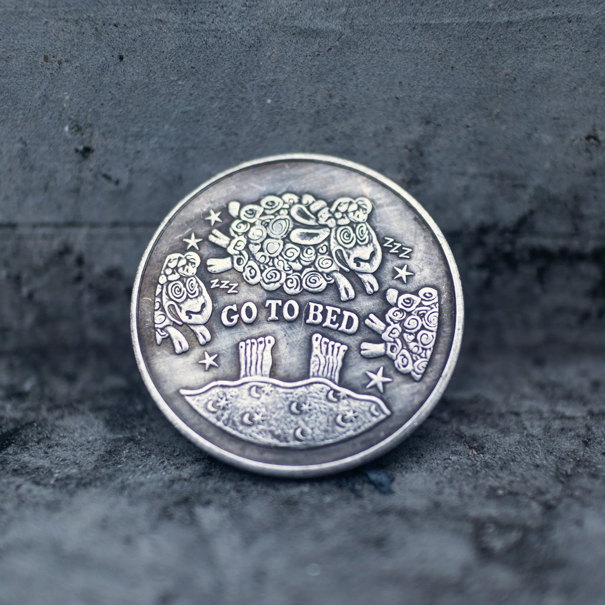Close-up of the Go To Bed face of a coin engraved with woolly sheep jumping over a moonlit hill, stars and ZZZ symbols, on dark stone surface