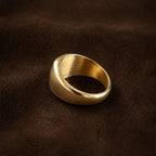A wide, chunky gold signet-style ring with a brushed and polished finish, resting on a dark brown textured leather surface.