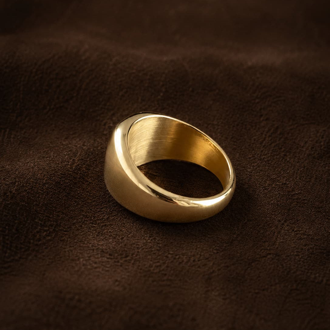 A wide, chunky gold signet-style ring with a brushed and polished finish, resting on a dark brown textured leather surface.