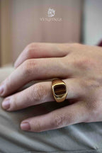 Gold tiger's eye ring worn on a pinky finger in natural light, showing the ring's rounded band profile and warm brown stone against a beige fabric