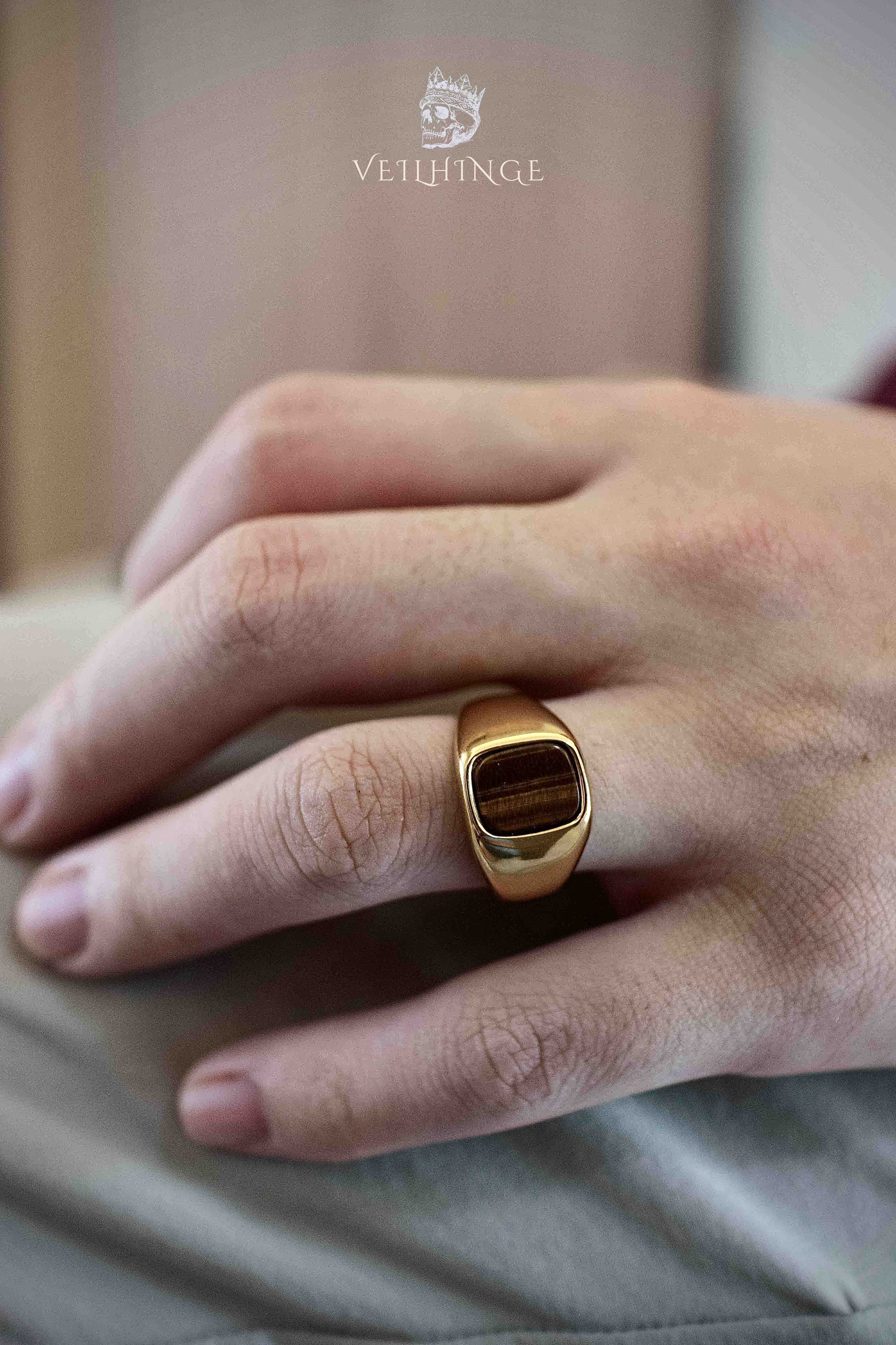 Gold tiger's eye ring worn on a pinky finger in natural light, showing the ring's rounded band profile and warm brown stone against a beige fabric
