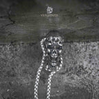 gothic skull heart pendant hanging on box chain against weathered stone wall — full drop view