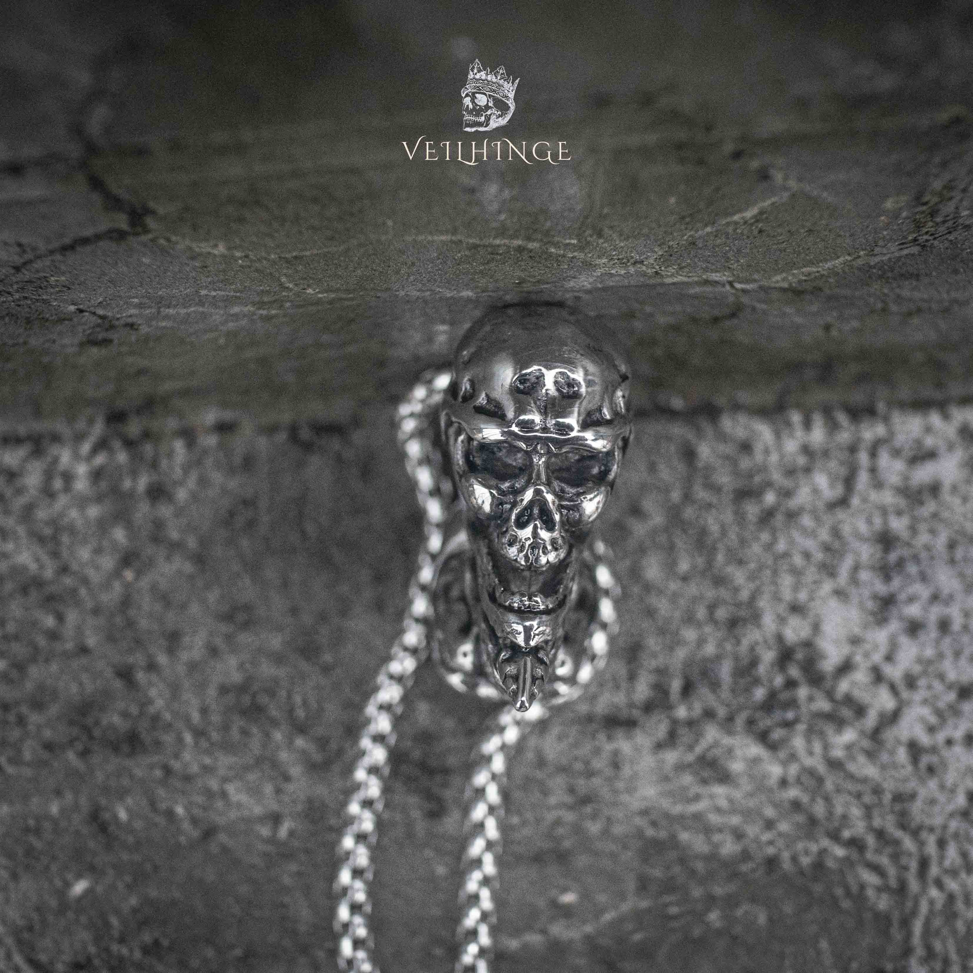 gothic skull heart pendant hanging on box chain against weathered stone wall — full drop view