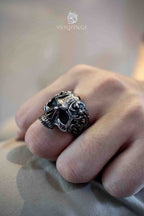 Gothic skull ring with baroque vine engravings worn on a clenched fist, deep hollow eye sockets and ornate floral scrollwork across the skull face in dark oxidized silver