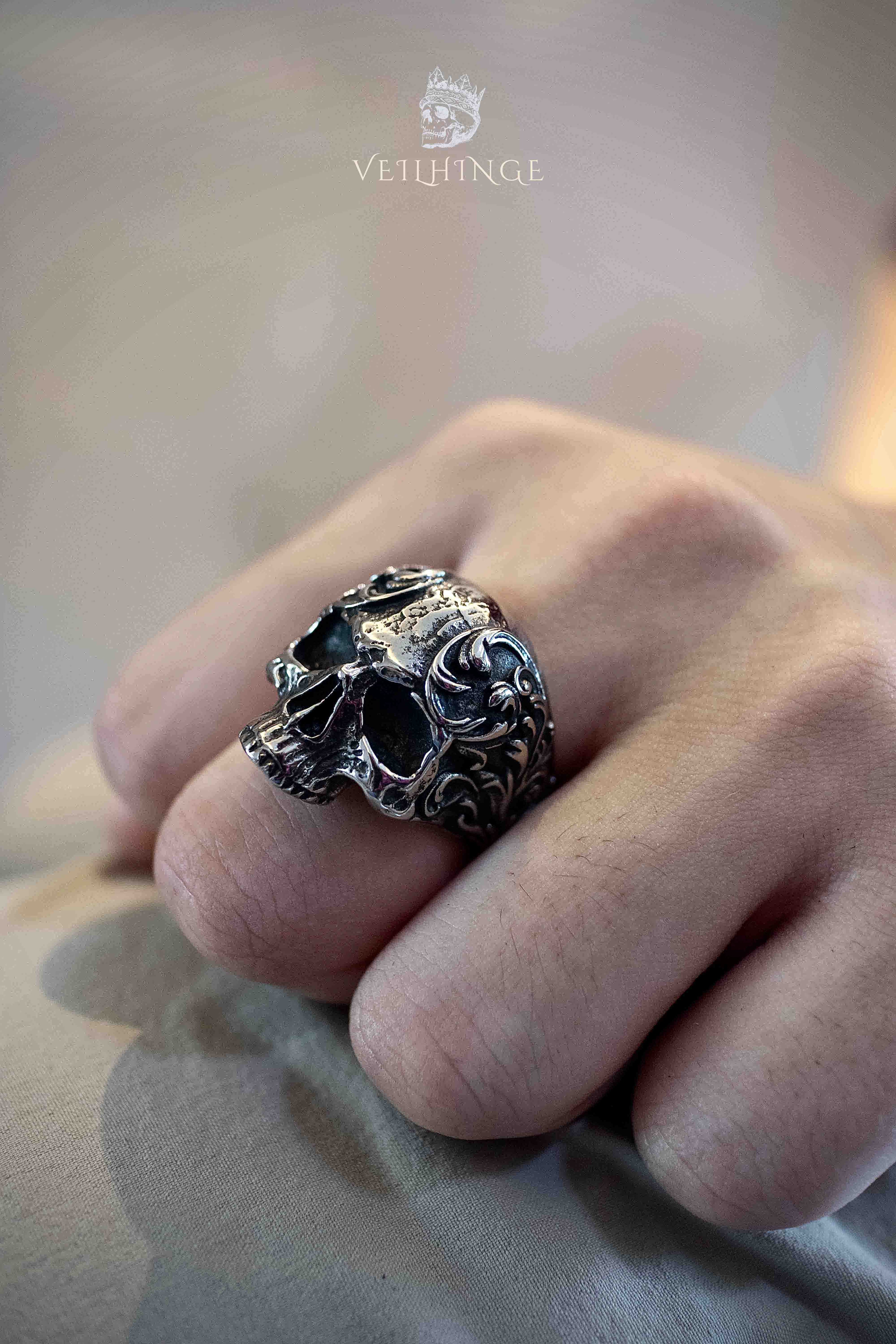 Gothic skull ring with baroque vine engravings worn on a clenched fist, deep hollow eye sockets and ornate floral scrollwork across the skull face in dark oxidized silver