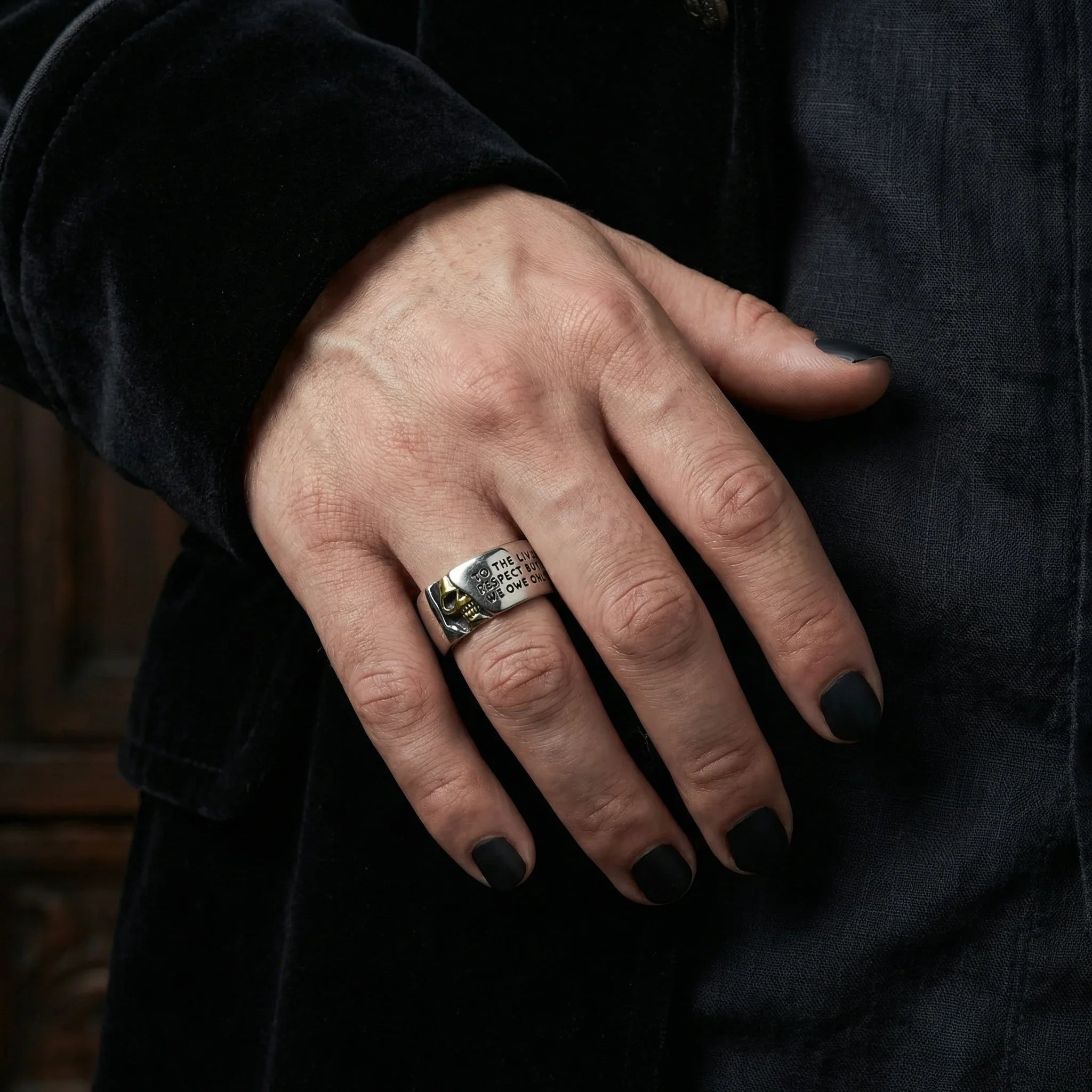 Hand in black velvet sleeve wearing the Gothic Voltaire skull ring with gold skull detail and engraved text