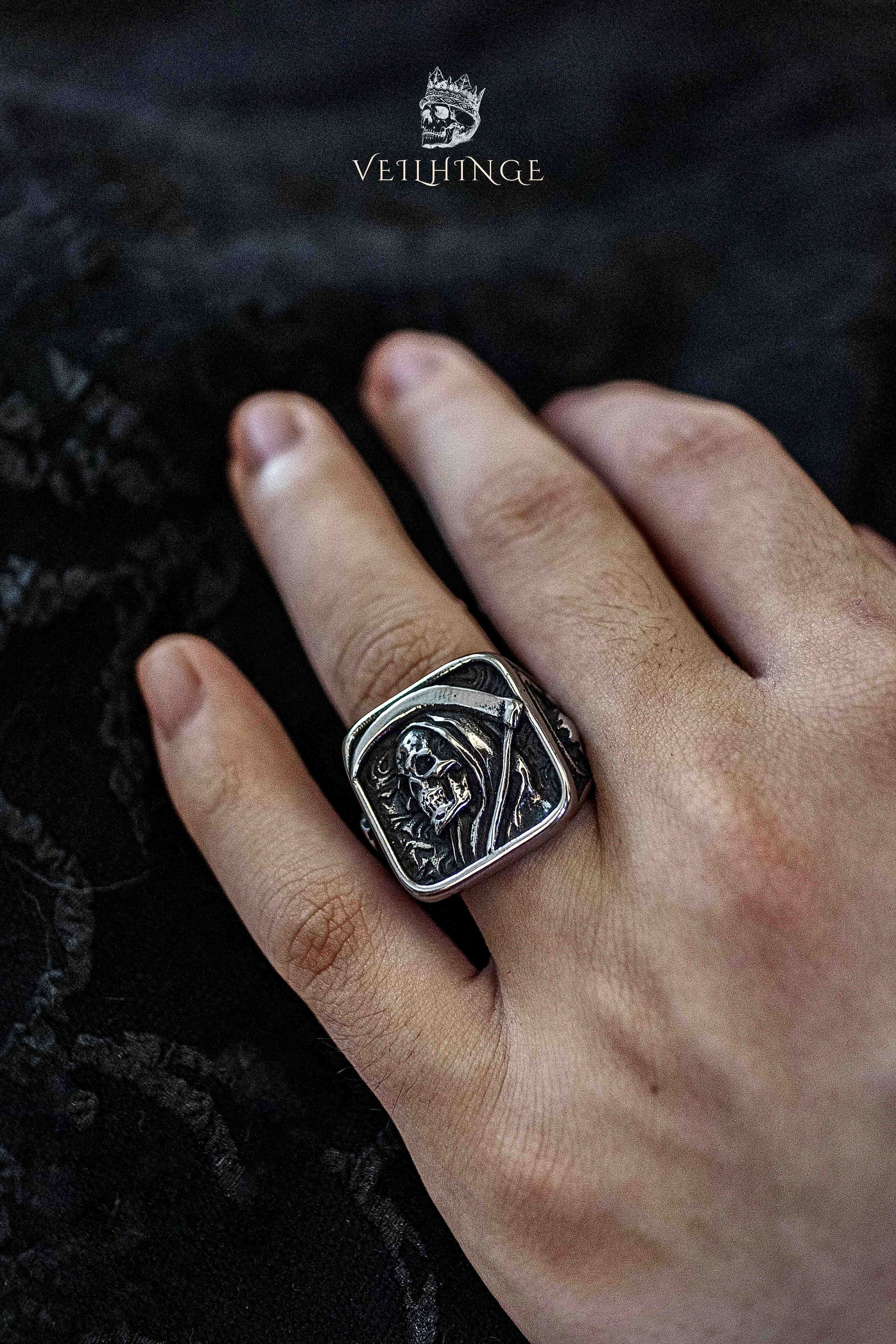 Grim Reaper hooded skull ring worn on a middle finger, deep relief engraving of a cloaked skeleton on a square oxidized silver face, against black velvet