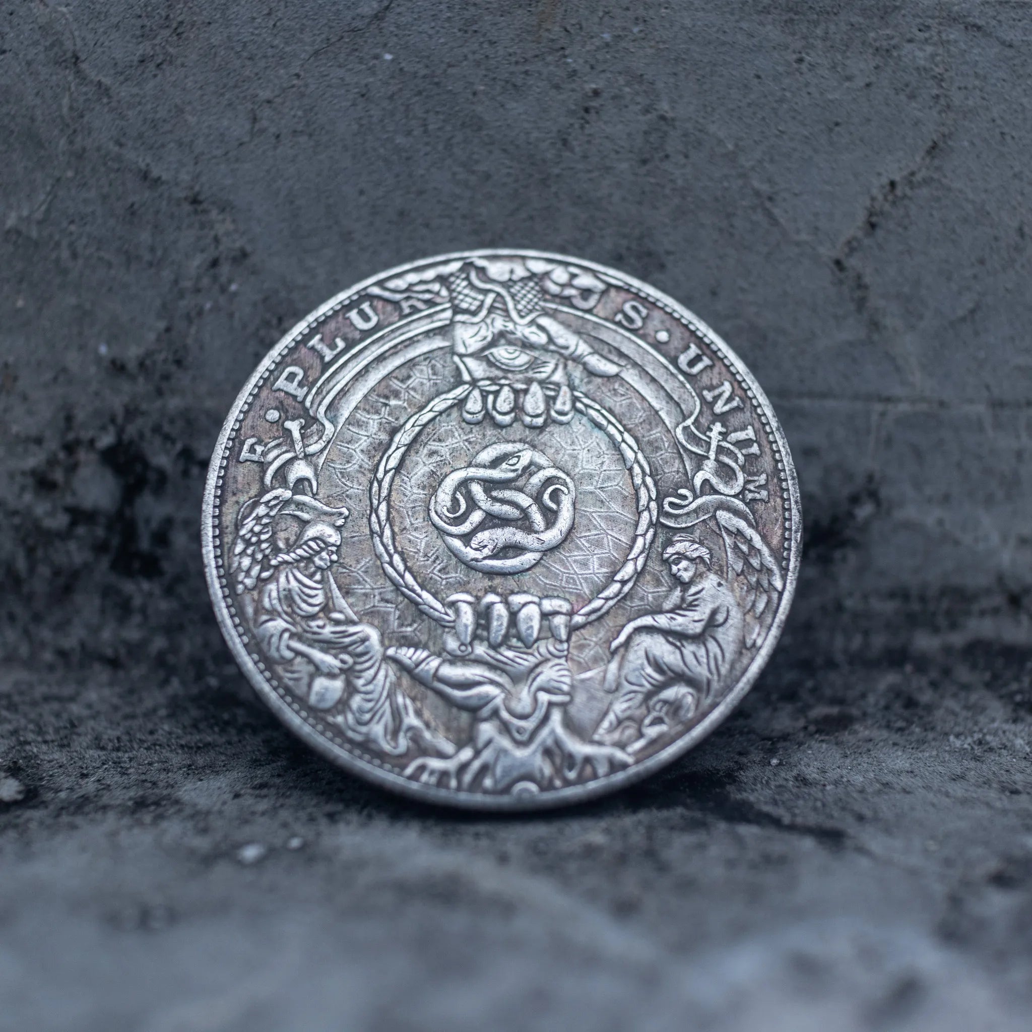 Close-up of a coin engraved with a coiled serpent encircled by a rope ring, two kneeling figures on either side, ornate crown detail above, E Pluribus Unum outer border