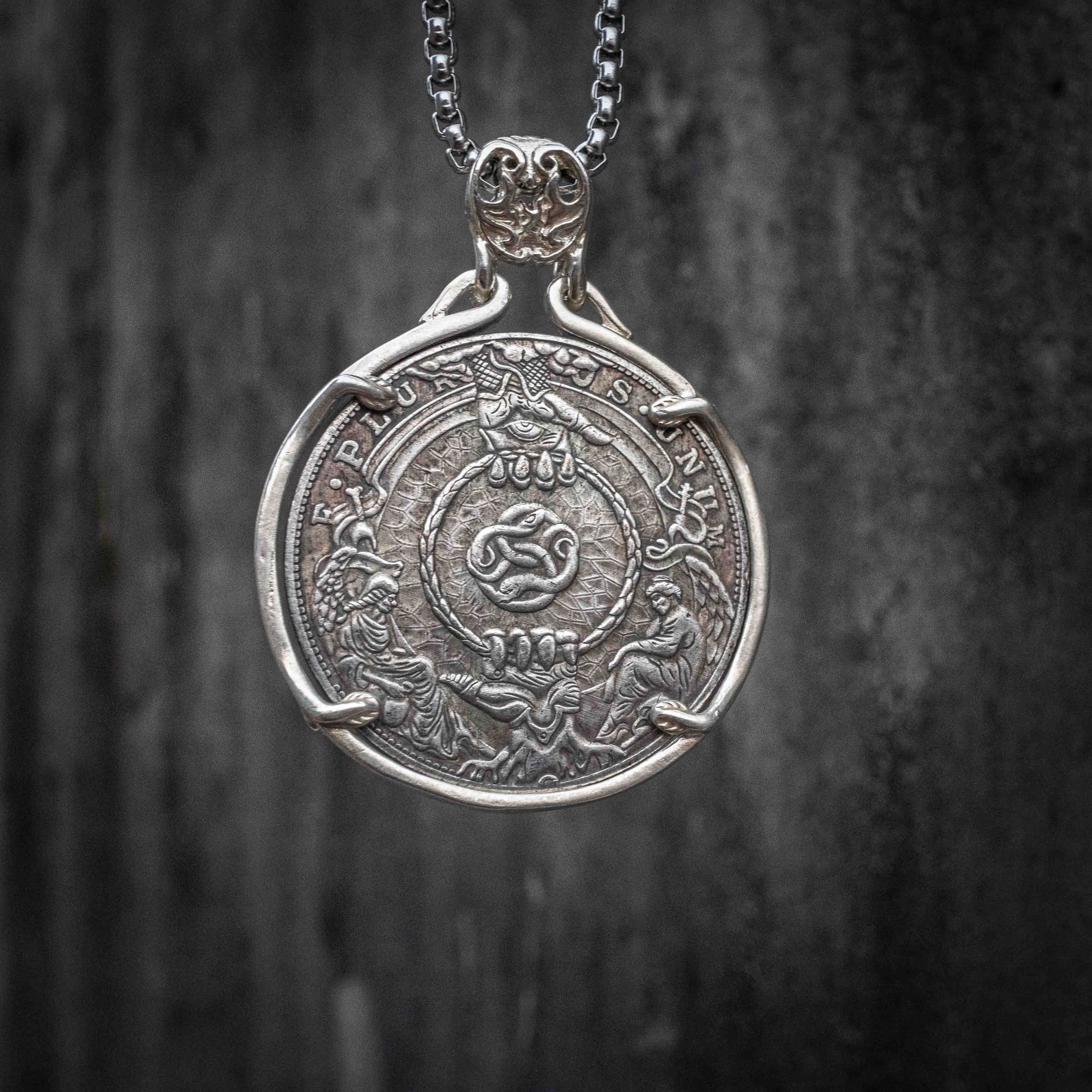 Coin pendant engraved with a coiled serpent at center framed by a rope ring, flanked by two kneeling figures holding the rope ends, E Pluribus Unum border, set in a four-prong bezel on a chain Main image