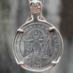 A silver pendant necklace featuring an antique-style religious coin medallion with a raised figure and the text 'MURIBLE LUXUM' and '1899', suspended from a silver chain against a blurred gray background.