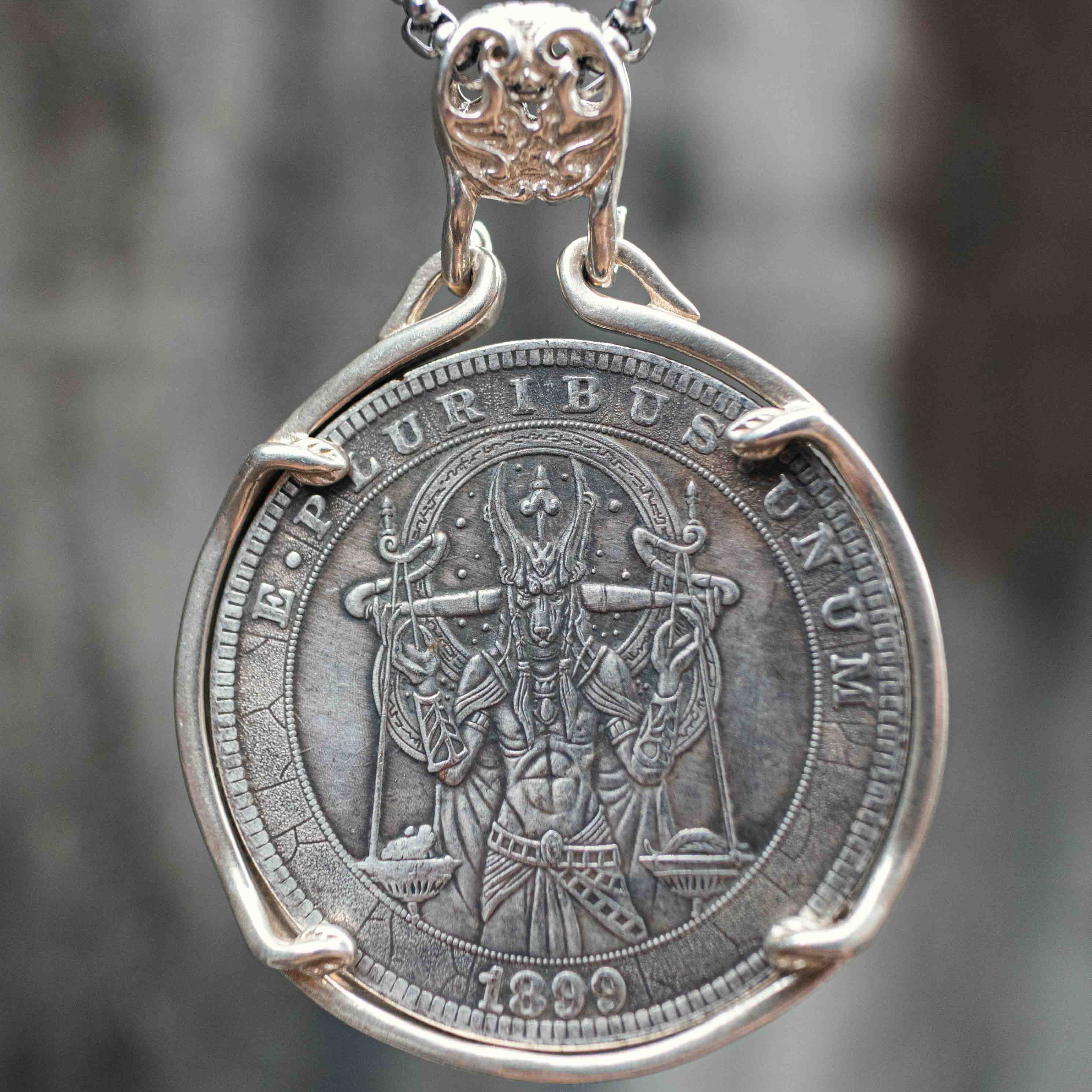 A silver pendant necklace featuring an antique-style religious coin medallion with a raised figure and the text 'MURIBLE LUXUM' and '1899', suspended from a silver chain against a blurred gray background.