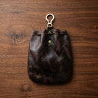 Back view of dark brown genuine leather drawstring pouch on walnut wood surface, showing natural patina and brass O-ring clip