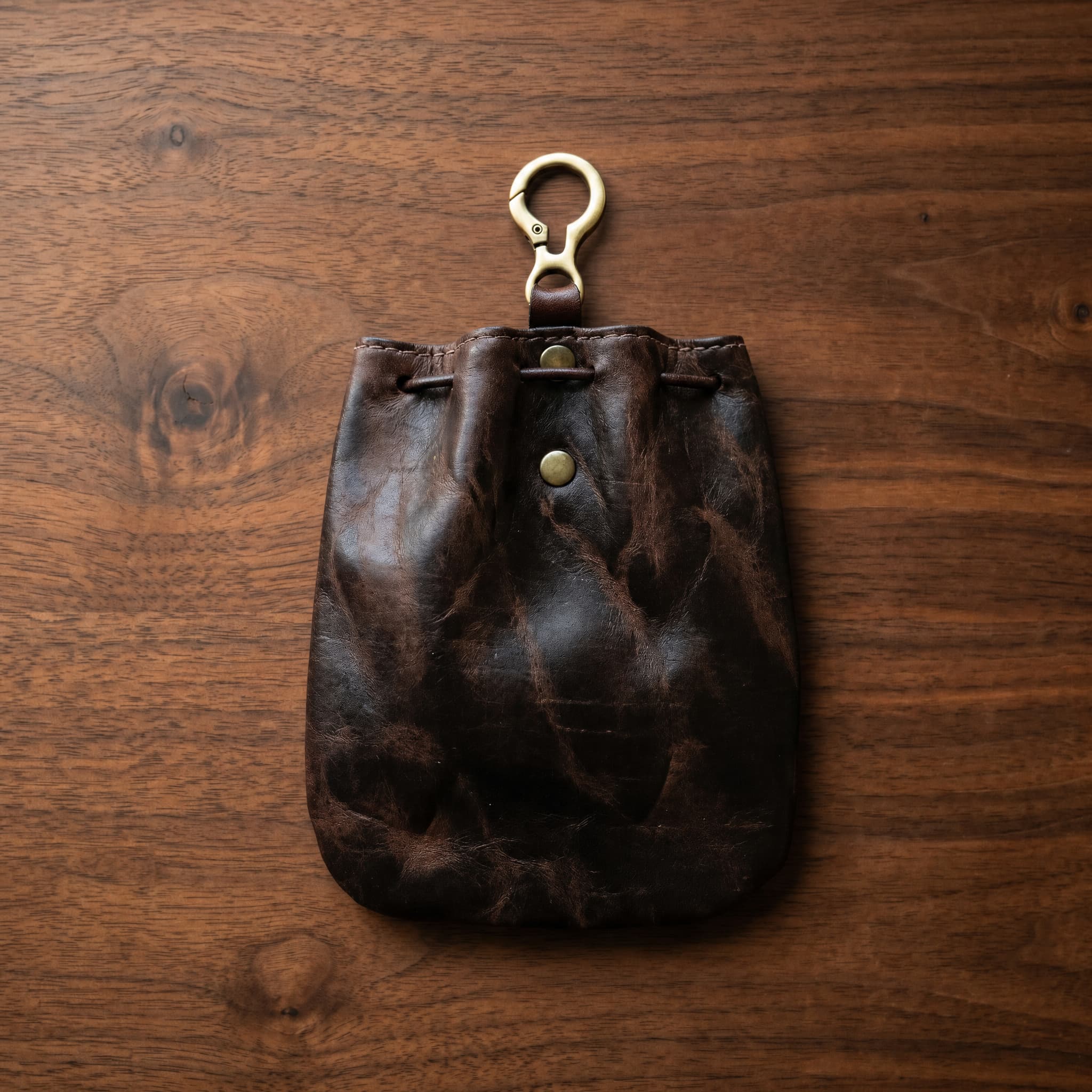 Back view of dark brown genuine leather drawstring pouch on walnut wood surface, showing natural patina and brass O-ring clip