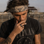 Man wearing a cream and navy longhorn skull paisley bandana as a headband, dressed in a vintage Nirvana tee with tattoos and rings, photographed at a ranch fence in golden hour light
