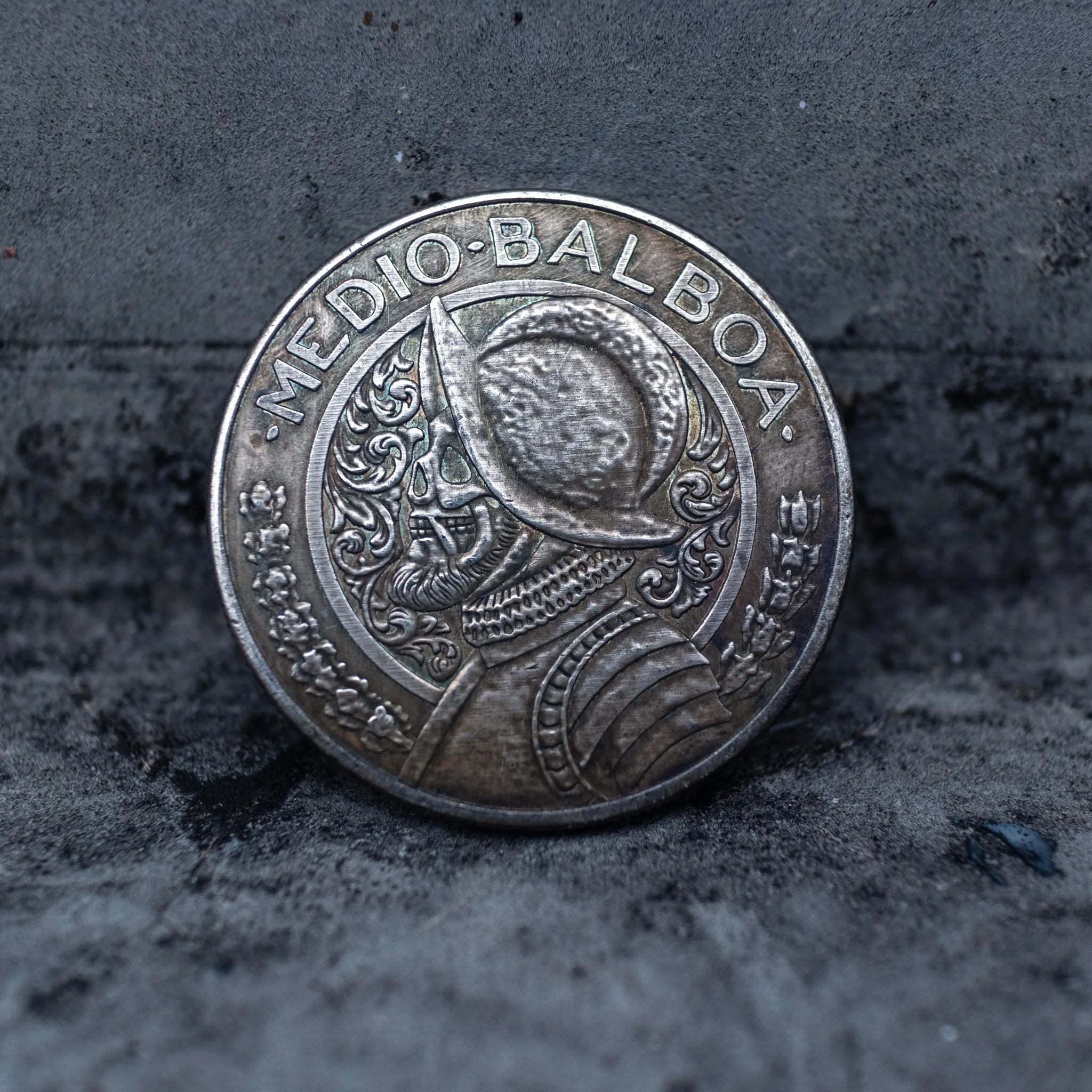Close-up of a coin engraved with a Spanish conquistador in a morion helmet and armor, ornate scrollwork background, Medio Balboa inscription across the top, on dark stone