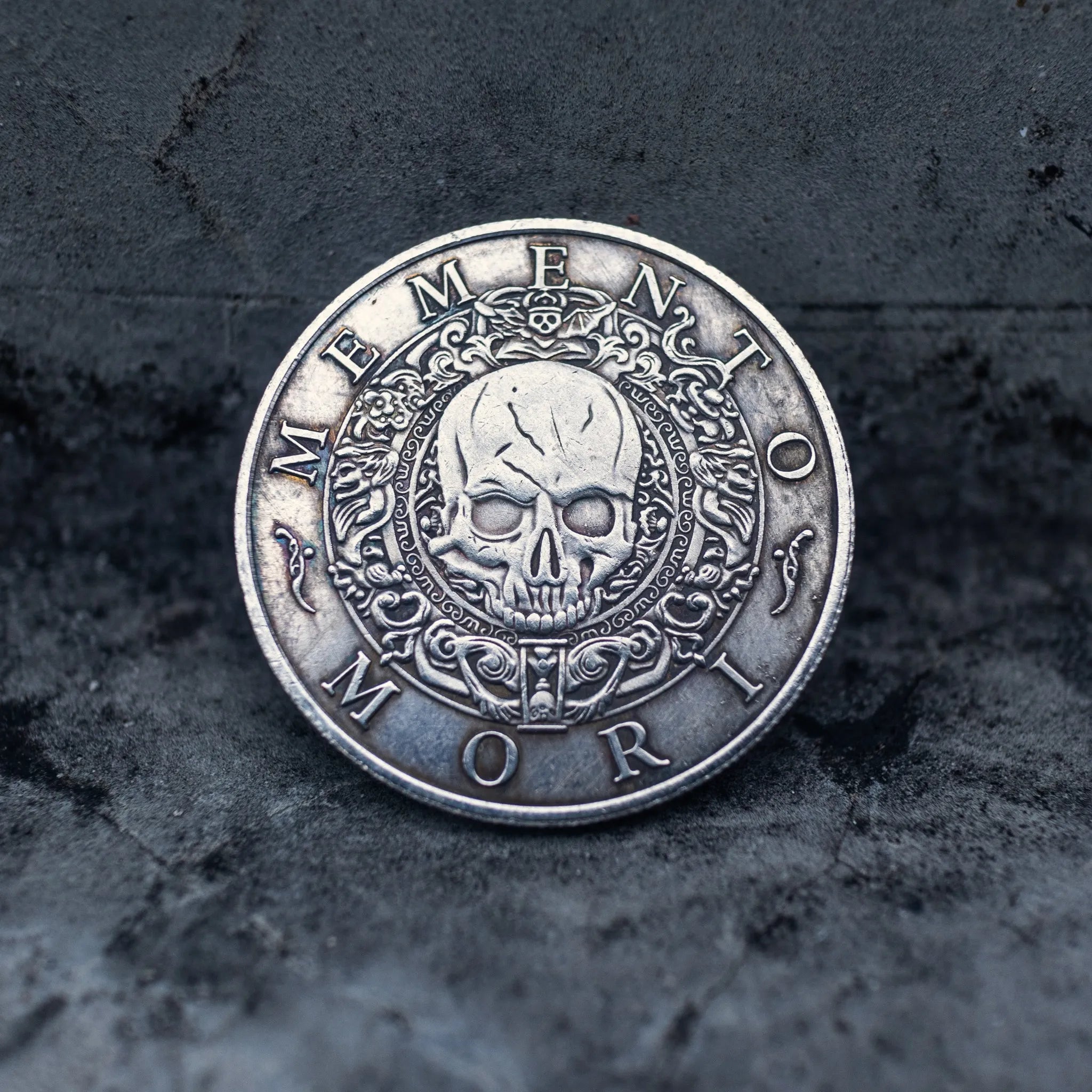 Memento Mori coin with ornate skull engraving and baroque border detail, displayed on dark textured stone