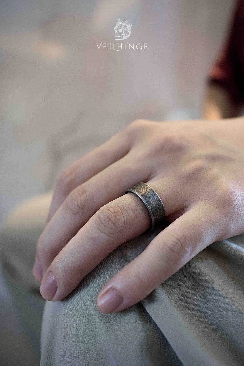 Minimalist wide plain band ring in brushed oxidized silver worn on a hand resting on fabric, smooth dark surface with a subtle hammered texture