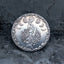 Close-up of a coin engraved with a flowing-haired goddess raising a full moon, crescent moon phases on either side, cloud and scrollwork border