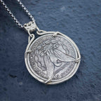 A silver pendant necklace featuring a coin-style medallion with an eagle design, suspended from a silver chain against a dark textured background.
