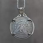 A silver pendant necklace featuring a coin-style medallion with an eagle design and a bail, suspended from a silver chain against a soft gray background.