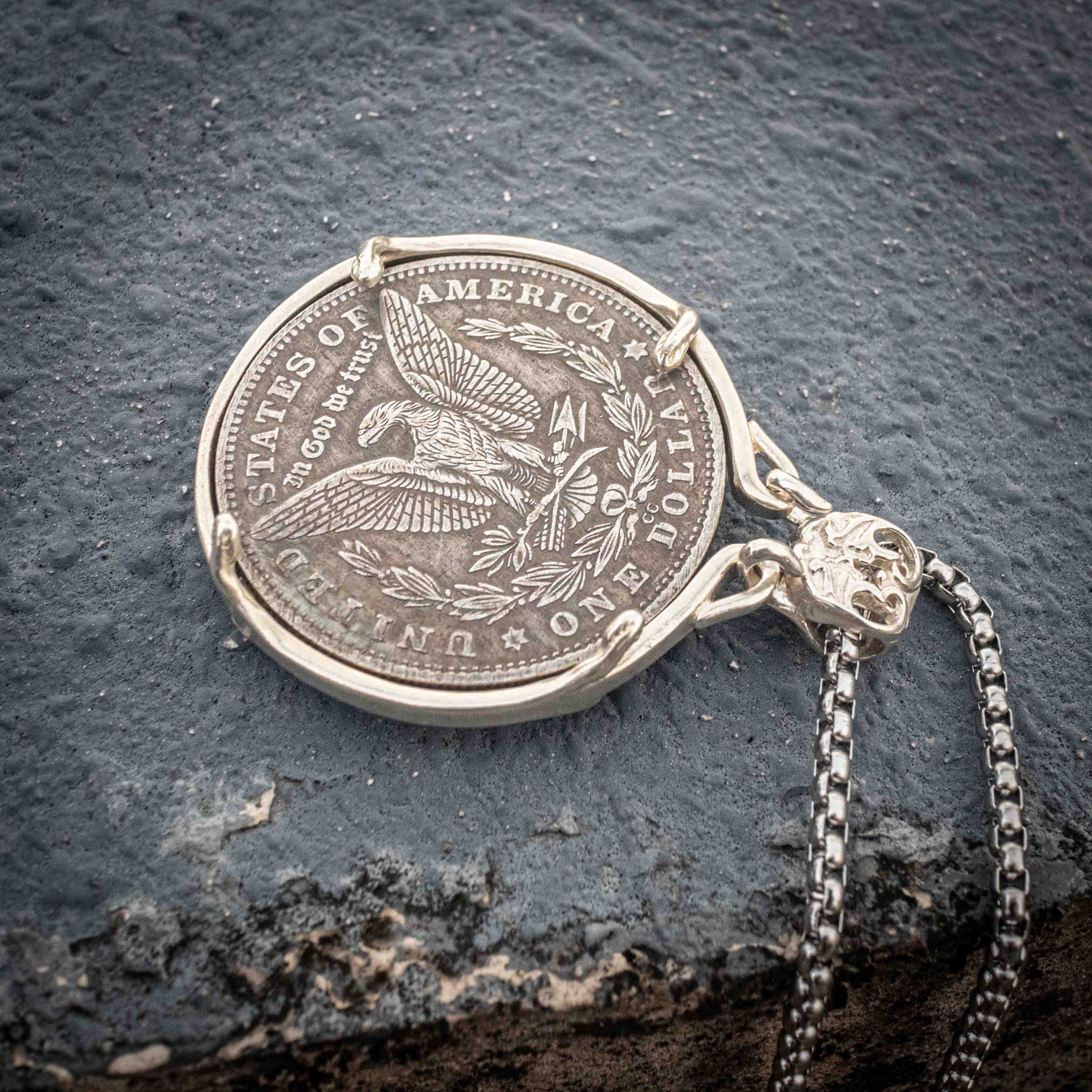 Heraldic serpent coin pendant laid flat on dark stone showing the Morgan Dollar eagle reverse with United States of America and One Dollar inscription