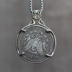 A silver pendant necklace featuring a coin-style medallion with an eagle design and engraved text, suspended from a silver chain against a blurred gray background.