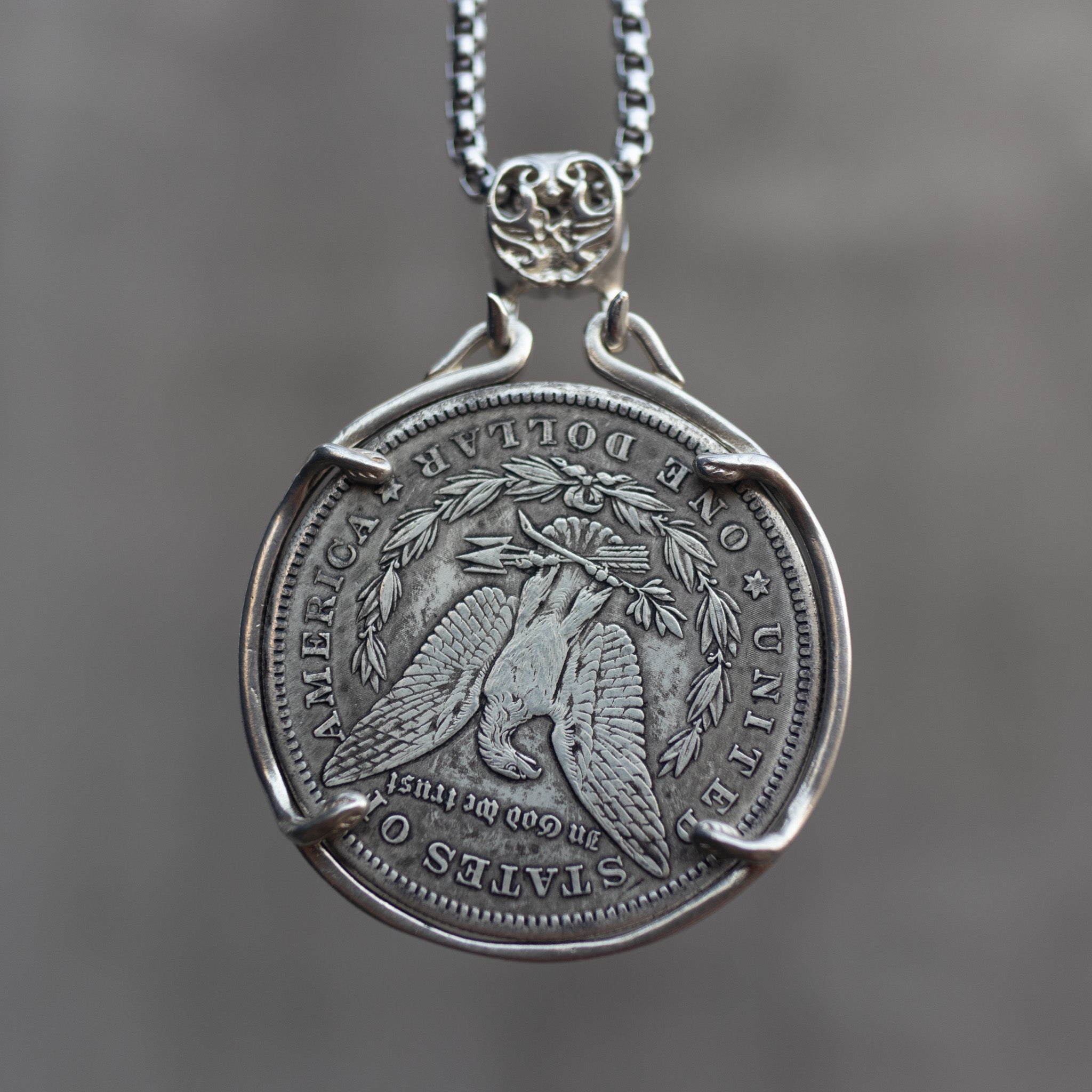 A silver pendant necklace featuring a coin-style medallion with an eagle design and engraved text, suspended from a silver chain against a blurred gray background.