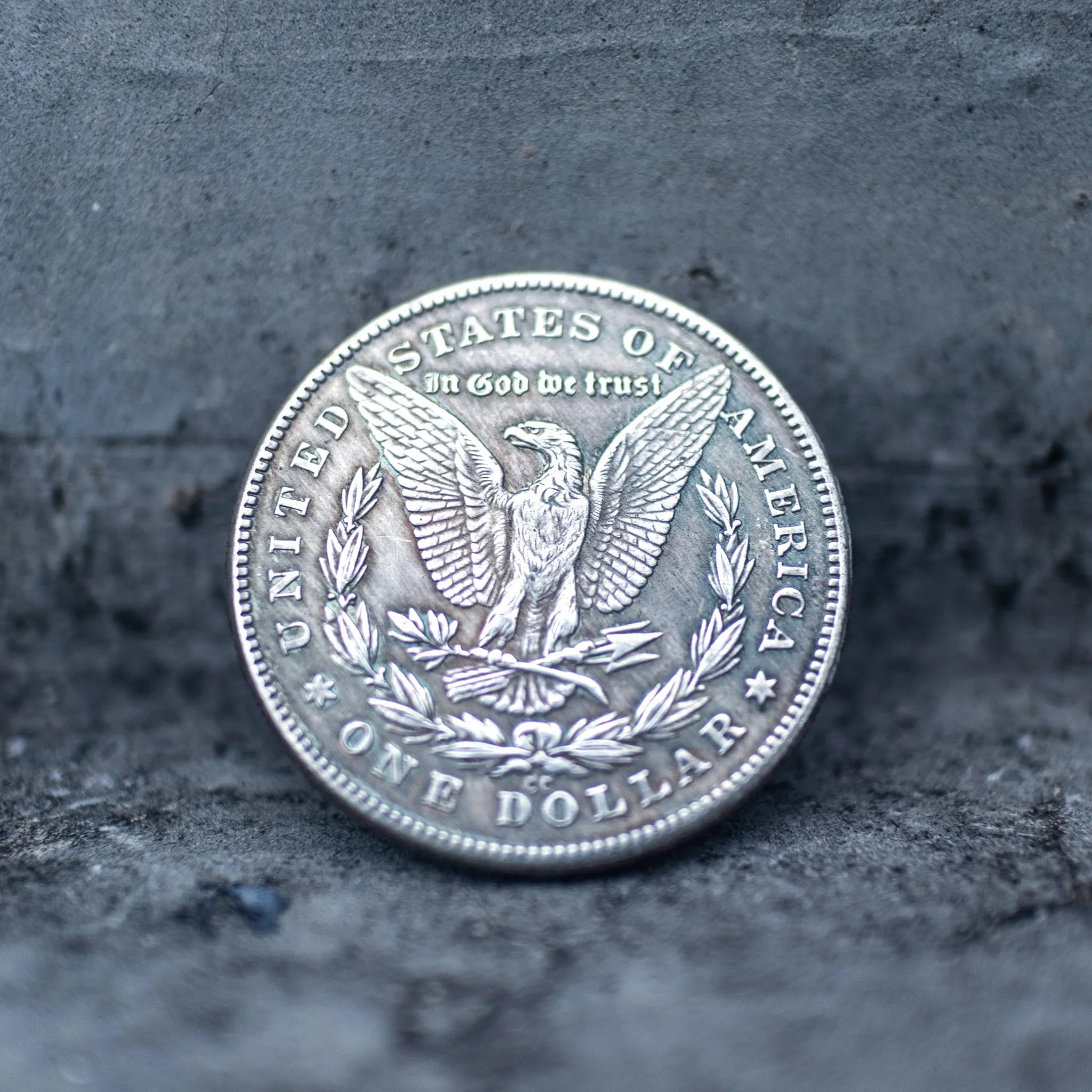 A silver-colored United States one dollar coin featuring an eagle and laurel wreath, photographed standing upright on a dark textured surface.