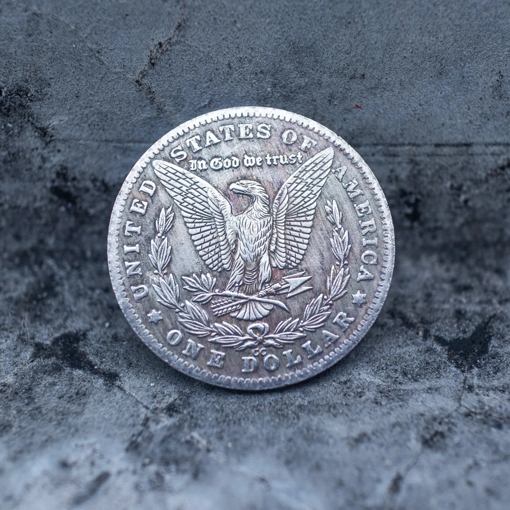 A circular silver-toned coin or medallion featuring an eagle and the text 'United States of America' and 'One Dollar,' photographed on a textured gray stone-like surface.