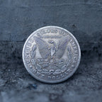 A silver-colored United States one dollar coin replica or pendant, featuring the eagle and 'In God we trust' inscription, resting on a textured dark gray surface.
