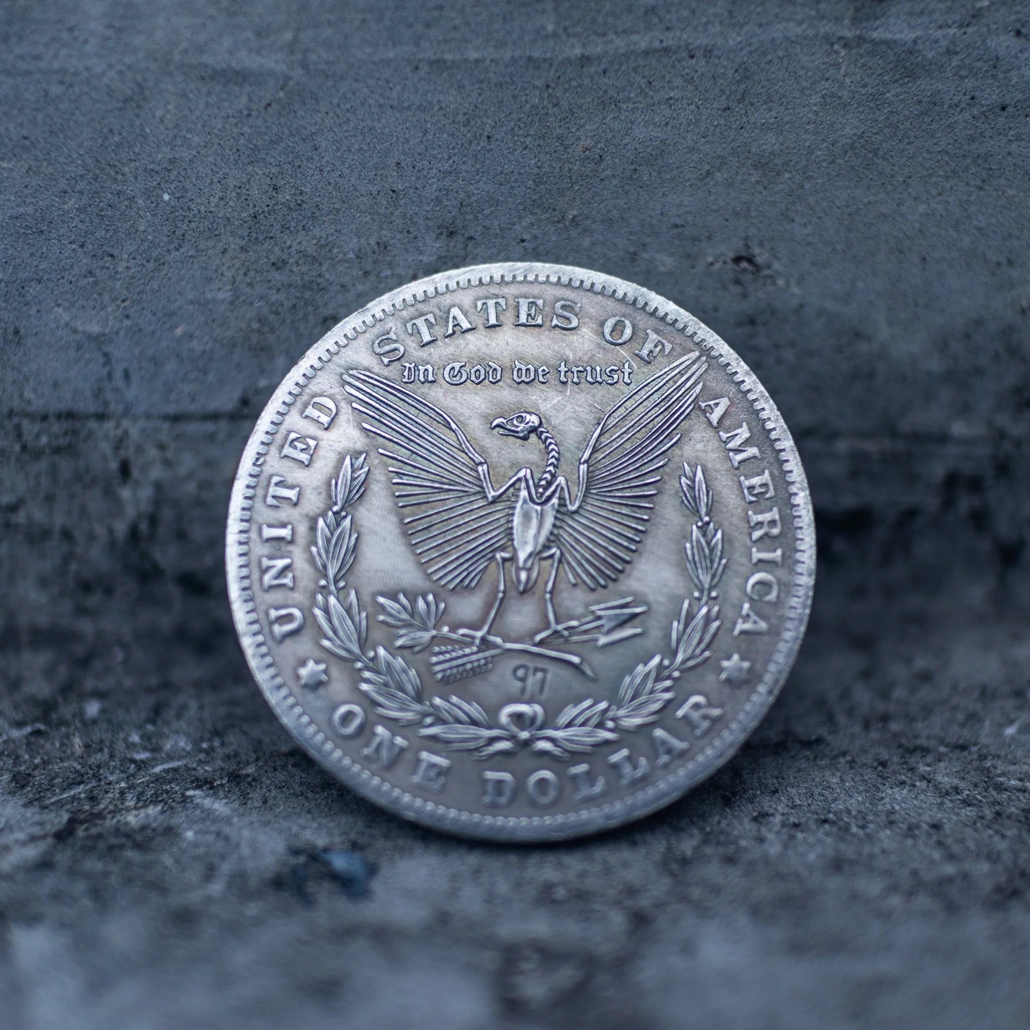 A silver-colored United States one dollar coin replica or pendant, featuring the eagle and 'In God we trust' inscription, resting on a textured dark gray surface.