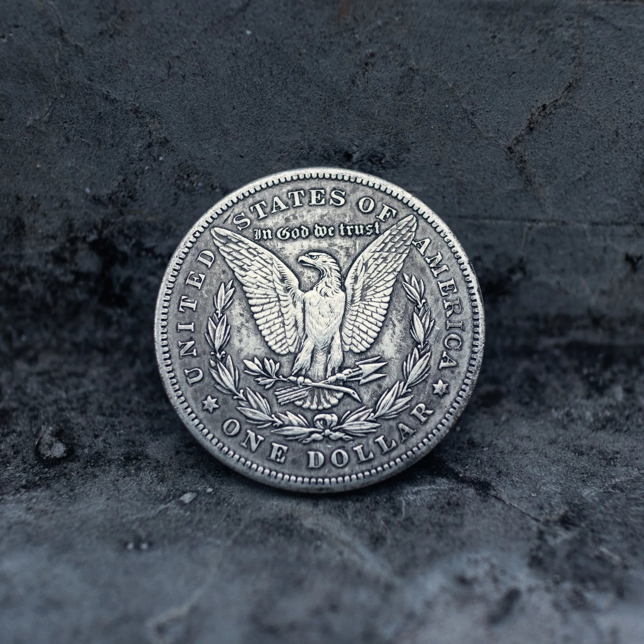 A silver-colored United States one dollar coin with the eagle design, photographed upright on a dark textured stone surface.