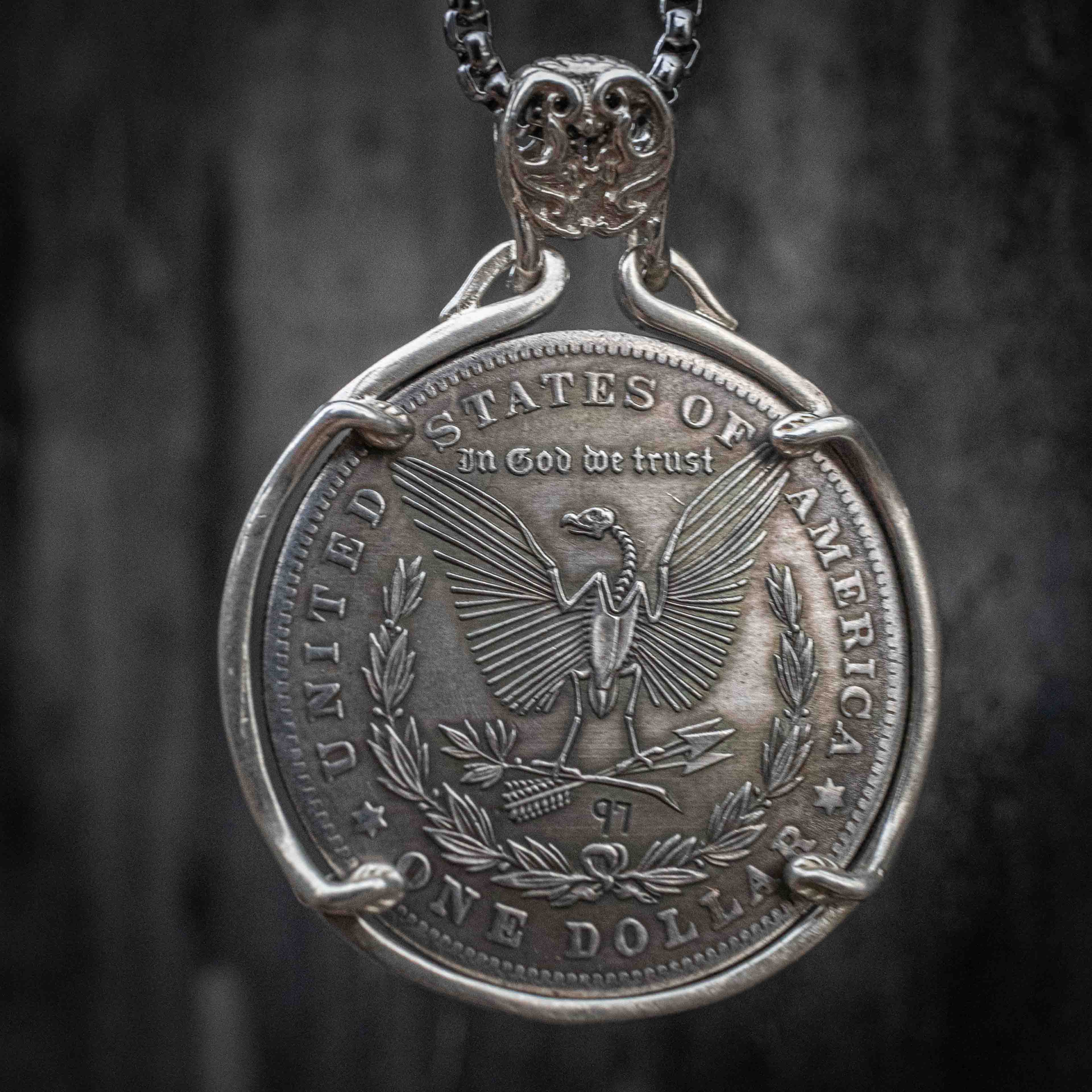 A silver pendant necklace featuring a coin-style medallion with an eagle and the text 'United States of America' and 'One Dollar,' suspended from a dark chain against a dark textured background.