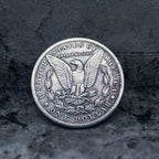 A silver-colored United States one dollar coin featuring an eagle and olive branch design, photographed from the reverse side against a dark textured stone background.