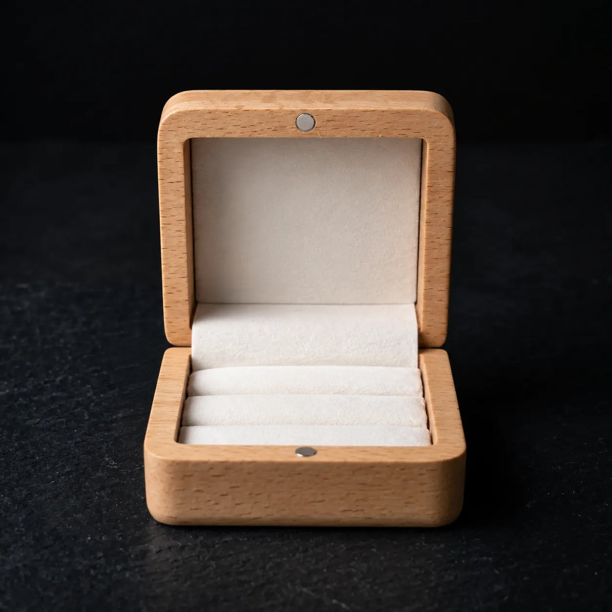 Open natural wood ring box with white velvet ring slots, shown from the front on a black background