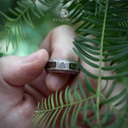 Norse rune ring held between fingers against pine foliage, transparent resin layer with gold-toned runic cutouts and Celtic knotwork border visible