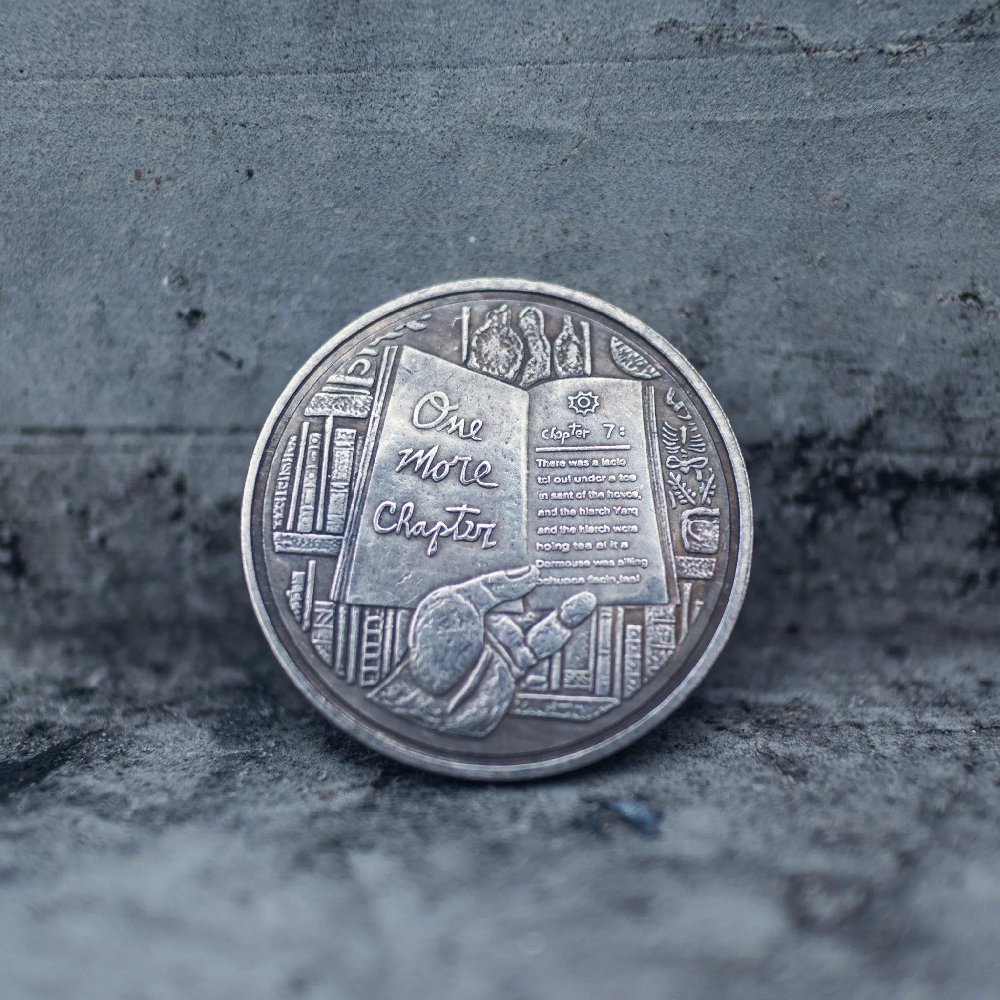 Close-up of the One More Chapter face of a coin engraved with a reader leaning against stacked books, open book and bookshelves in background