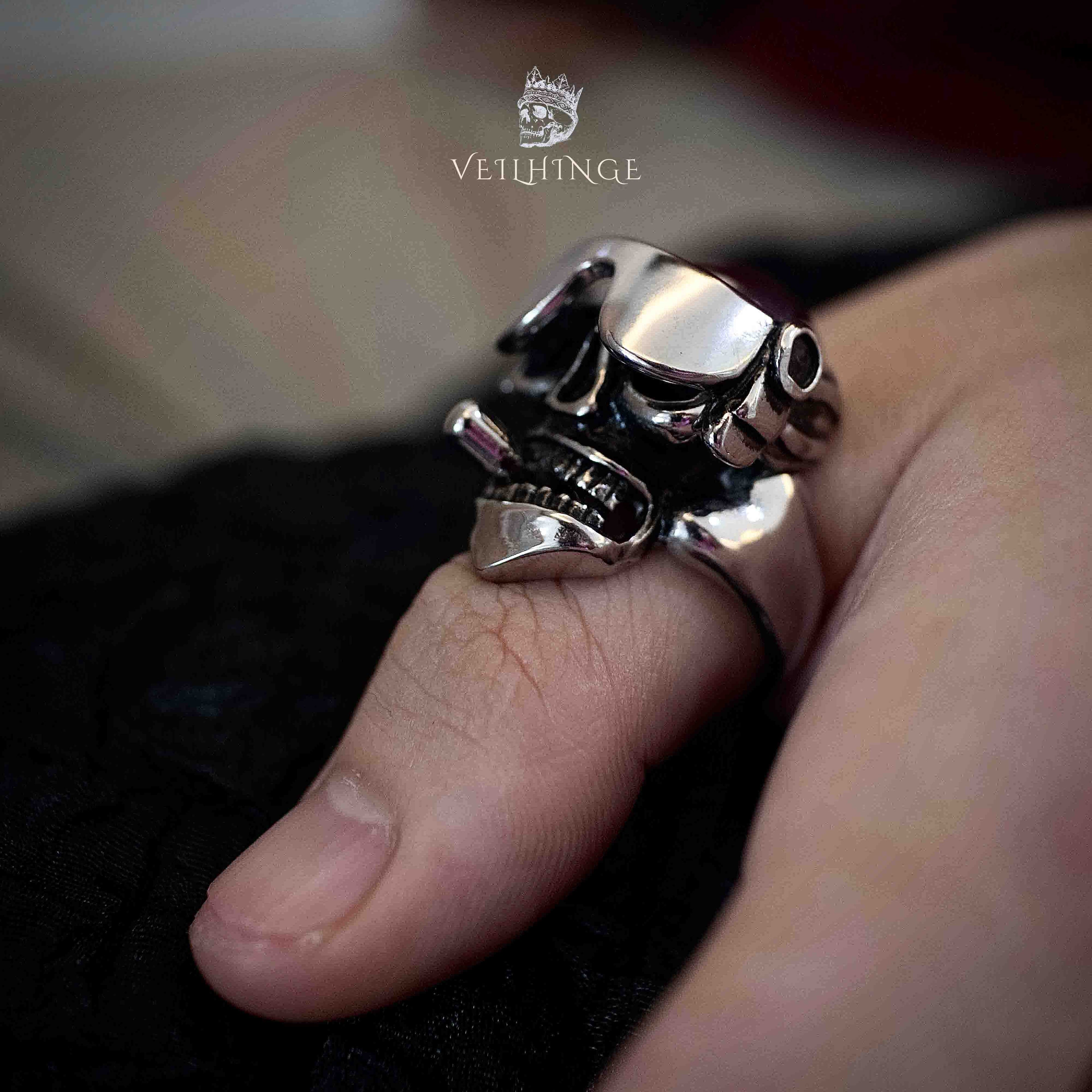 Oversized 3D skull ring in polished silver worn on a thumb, wide cheekbones and open jaw visible from a side angle, bold scale against a black fabric background