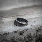 Oxidized black signet ring angled view showing blank rectangular face and rounded sandblasted stainless steel band, minimalist dark aesthetic ring by Veilhinge