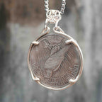 A silver pendant necklace featuring a circular coin charm with an eagle design, suspended from a silver box chain against a blurred gray and brown background.