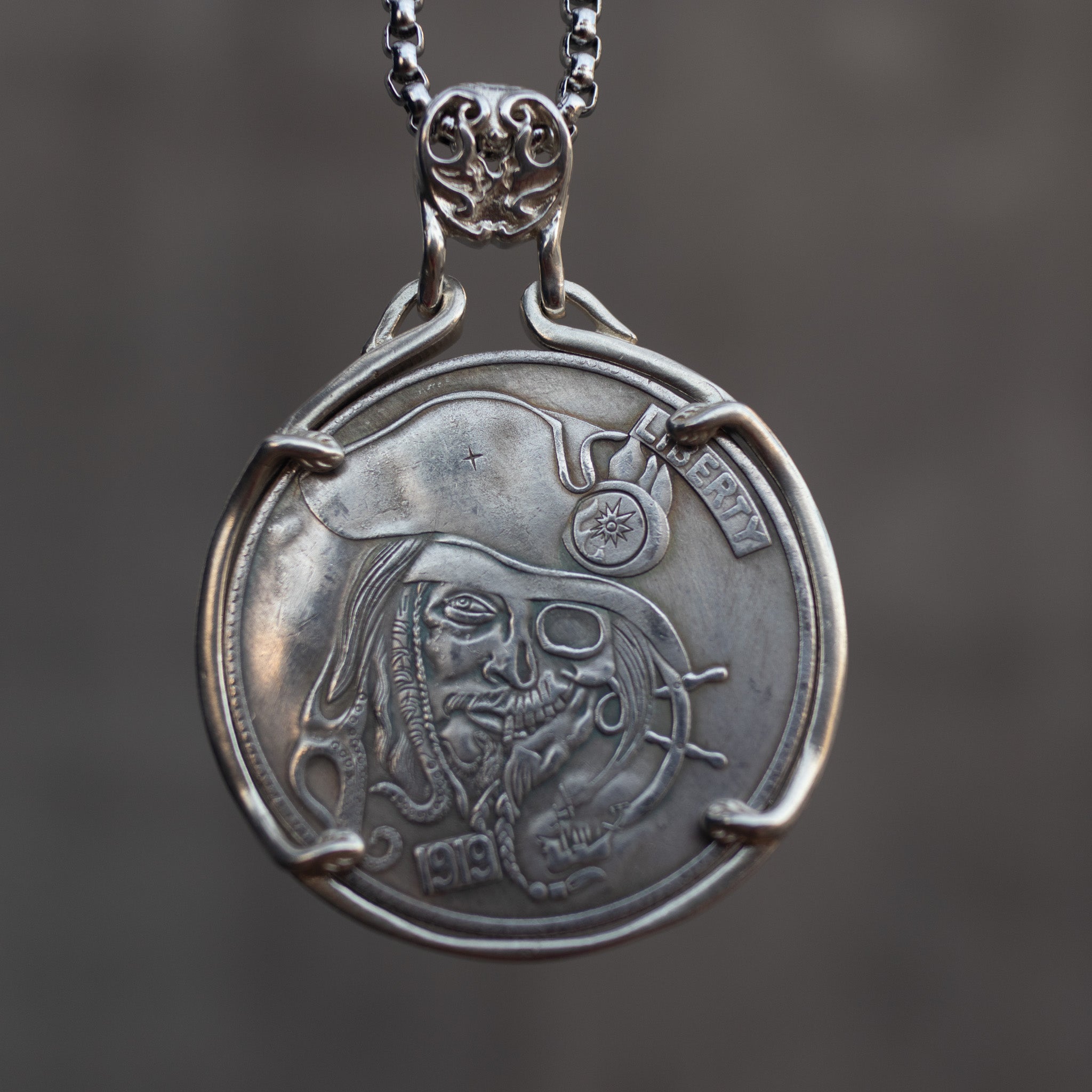 Coin pendant engraved with a bearded pirate captain skull face in tricorn hat with compass badge, ship wheel detail, set in a four-prong bezel on a box chain Main image