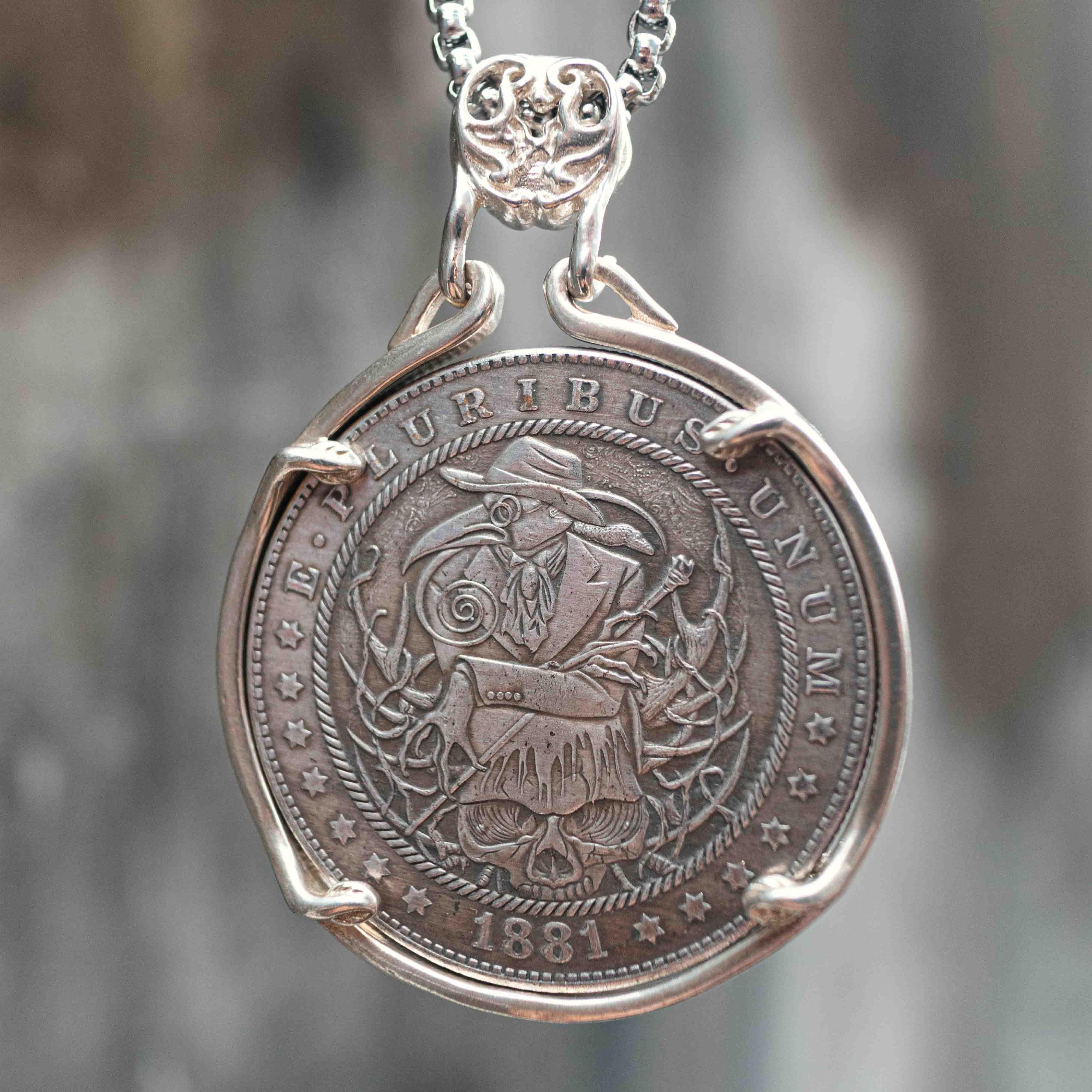 Coin pendant engraved with a plague doctor in a beak mask and long robe surrounded by skull and vine details, dated 1881, set in a four-prong bezel on a chain Main image