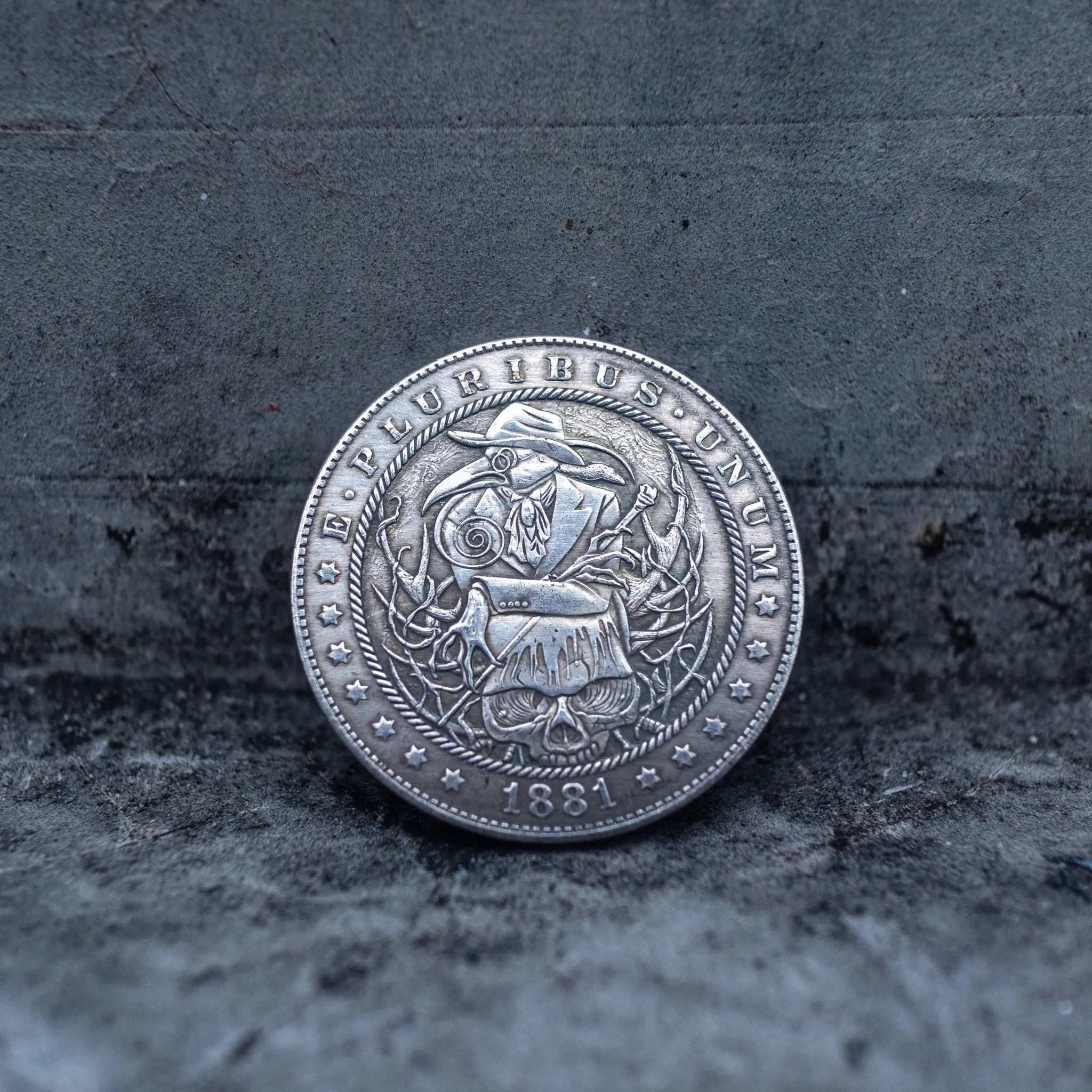 Close-up of a coin engraved with a plague doctor in a beak mask and robes, skull and scrolling vine details, E Pluribus Unum border, dated 1881, on dark stone