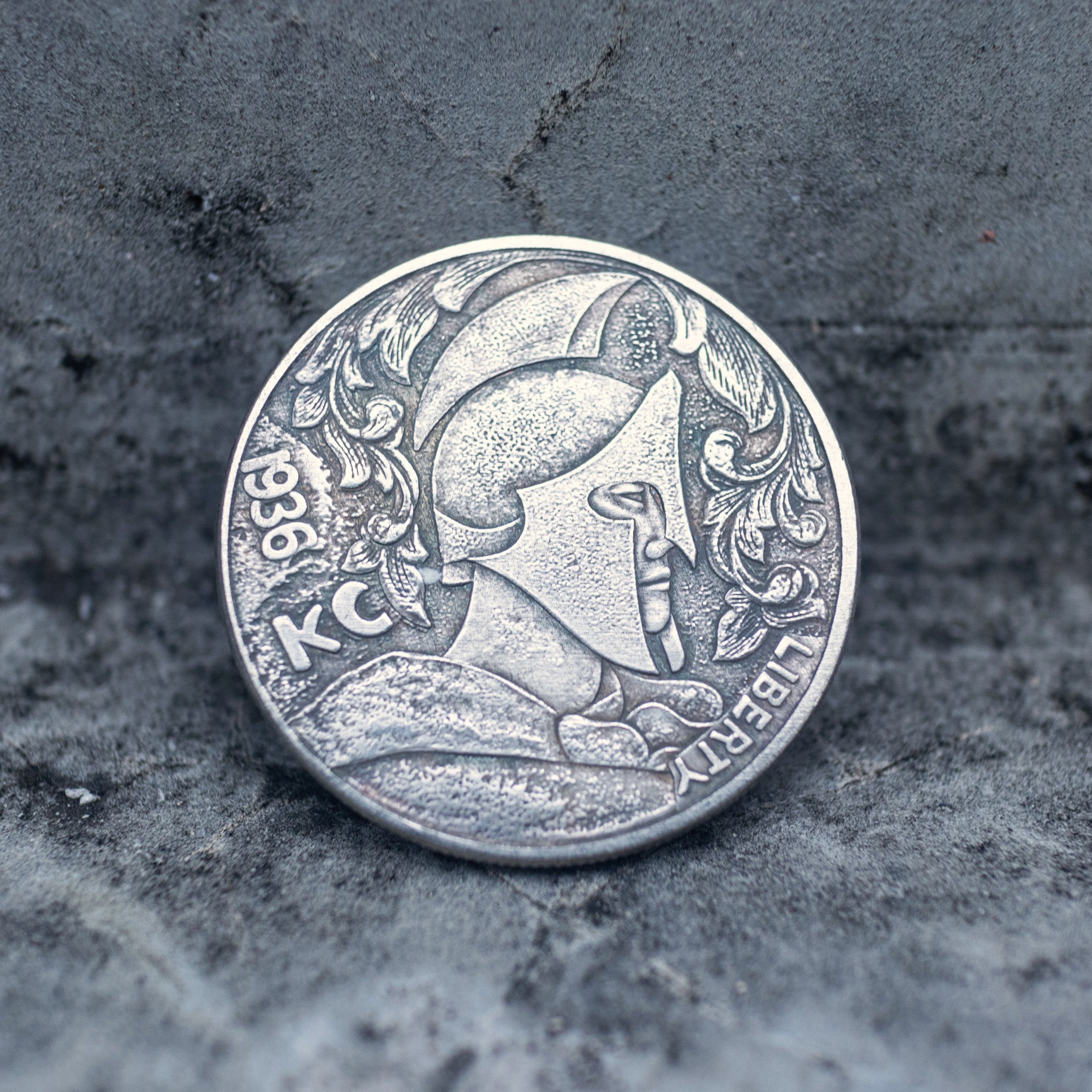 Close-up of a coin engraved with a Roman centurion profile in a Corinthian helmet with nose guard and decorative foliage crest, KC mark, Liberty inscription, dated 1936, on dark stone