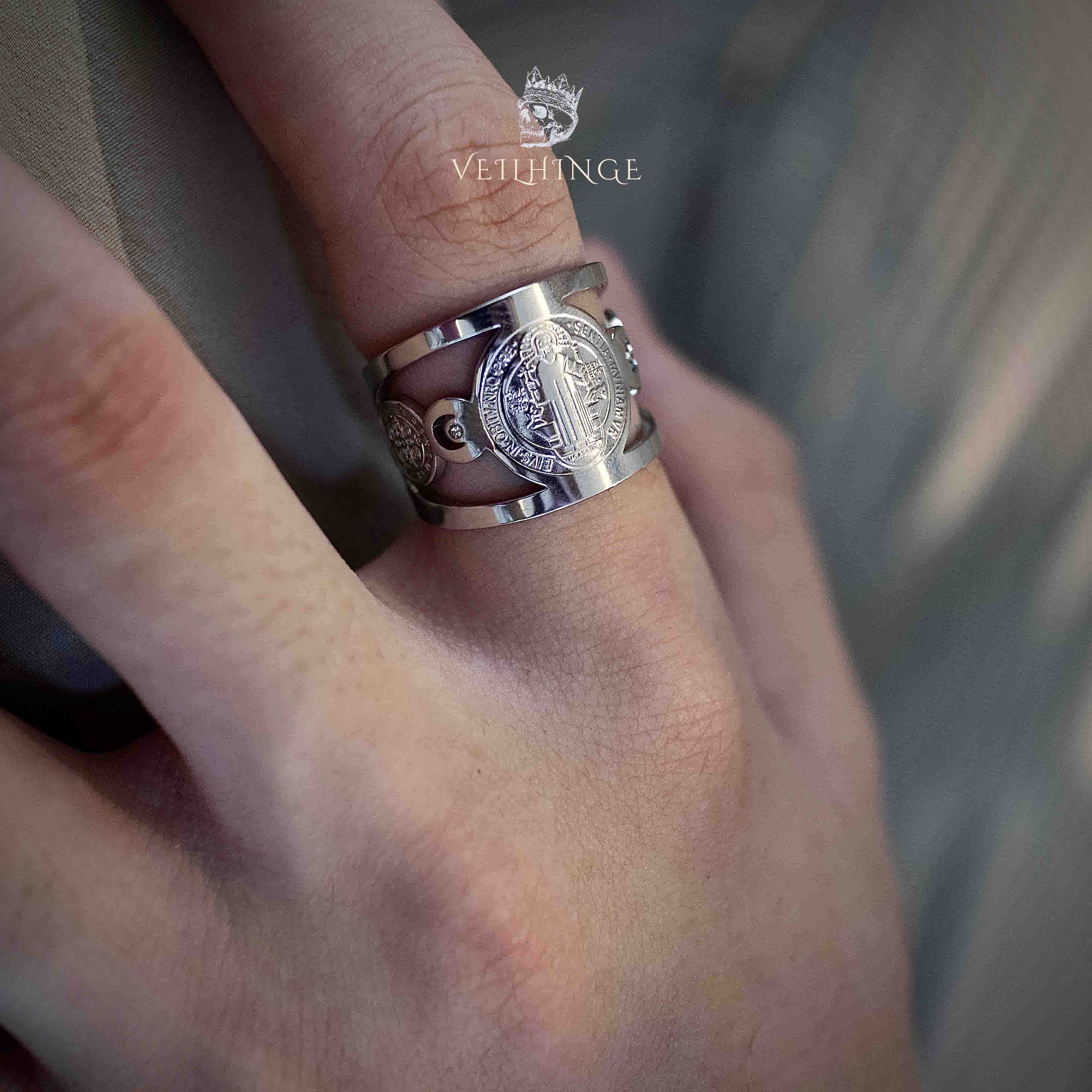 Saint Benedict medal ring worn on a finger, wide silver band with cutout scrollwork on the sides and oval medal face visible from a side angle