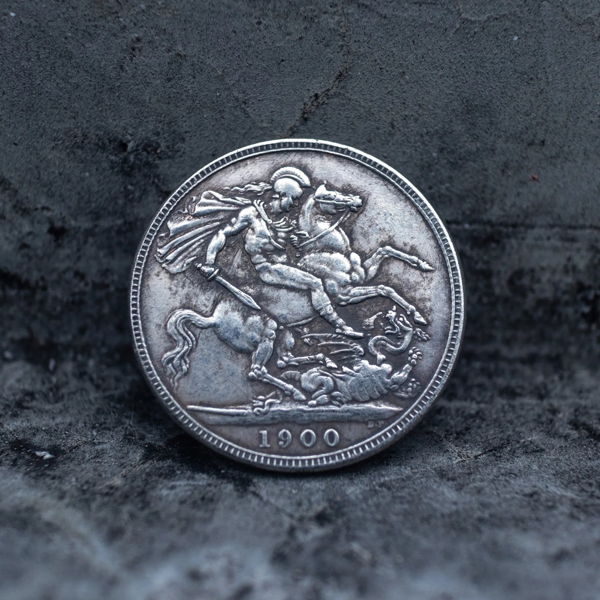Close-up of a coin engraved with Saint George on a rearing horse driving a lance into a dragon below, dated 1900, on dark stone surface