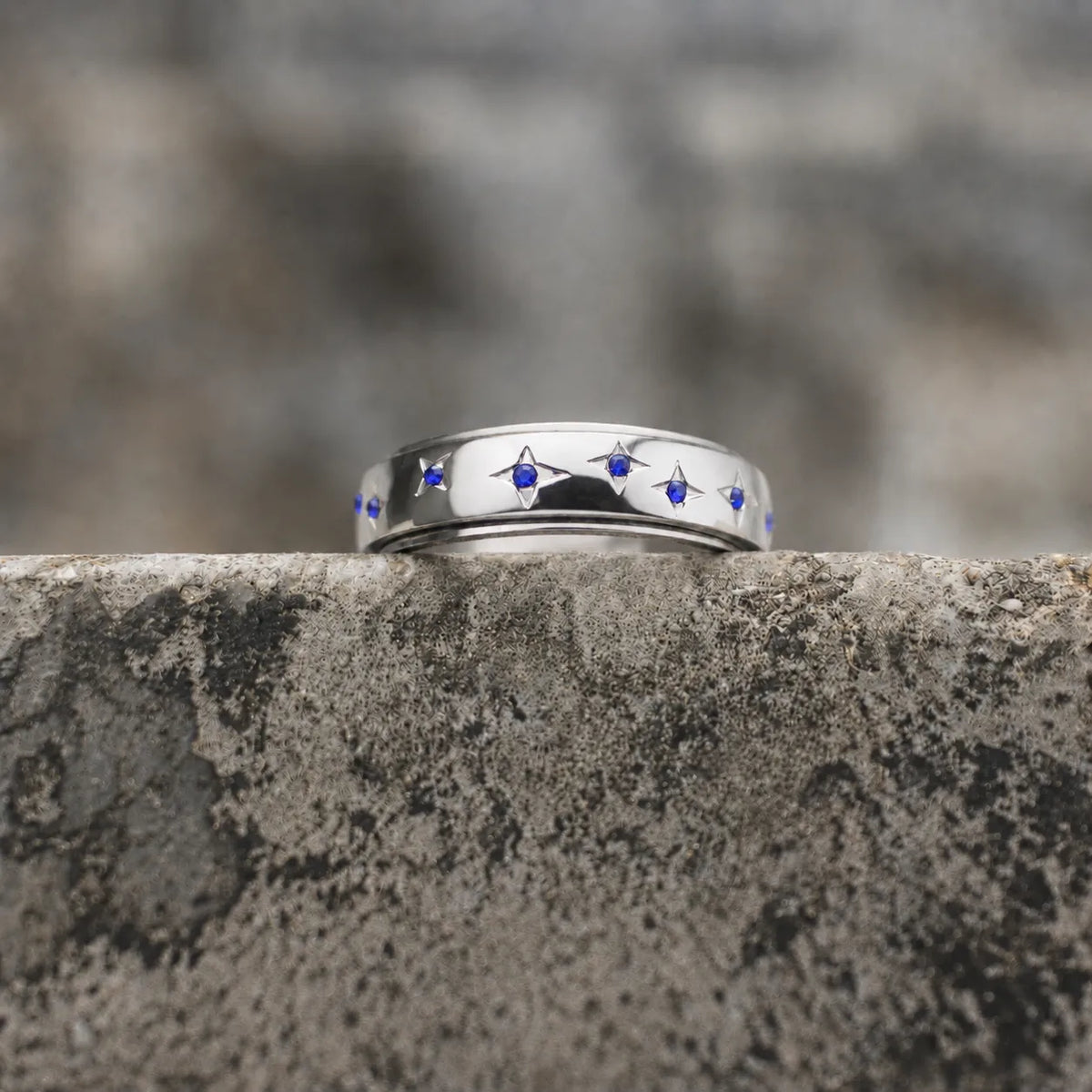 A polished silver ring featuring a row of small blue gemstone star motifs, resting on a textured gray stone ledge with a blurred natural background. Main image